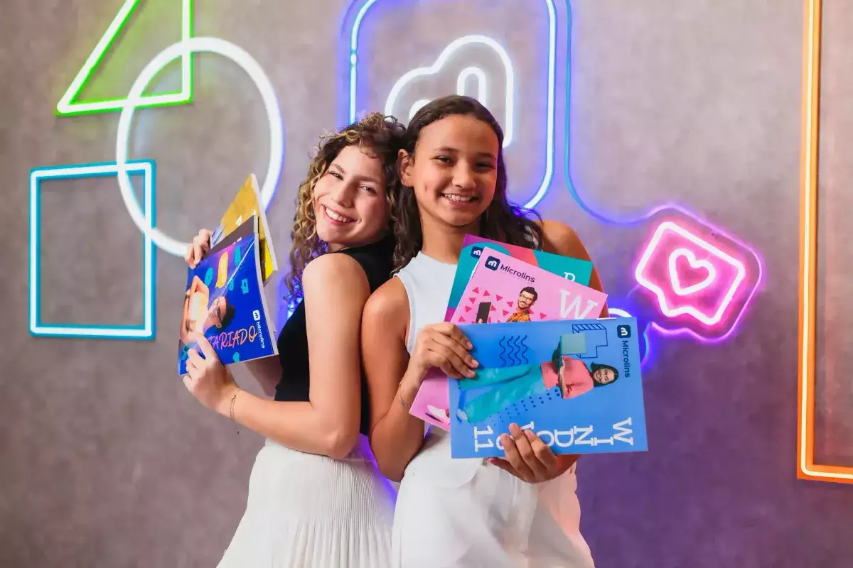 Duas alunas da Microlins sorrindo e mostrando apostilas do curso